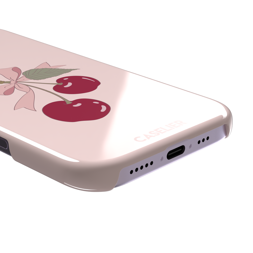 Soft pink phone case featuring two glossy red cherries tied with a pink ribbon bow and green leaf illustration in a coquette style, phone case by Caselier — snap case, charging port detail